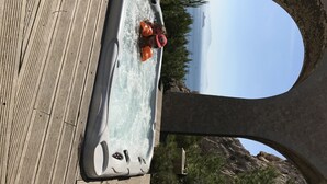 Un jacuzzi con dos personas, una terraza de madera y un puente de piedra al fondo.