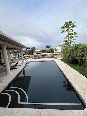 A swimming pool with a clear water surface, surrounded by a paved deck and a covered seating area.