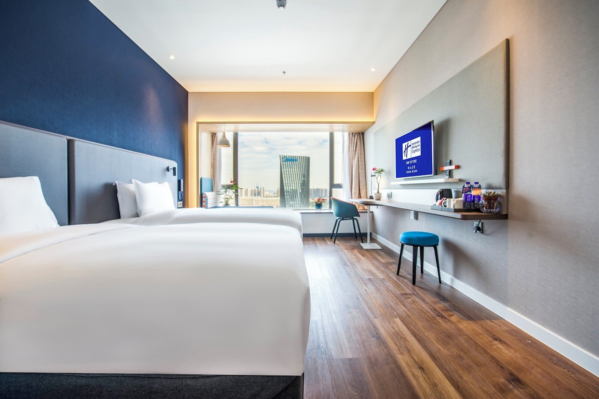 A hotel room with a large bed, a desk, a chair, a TV, and a view of a cityscape.
