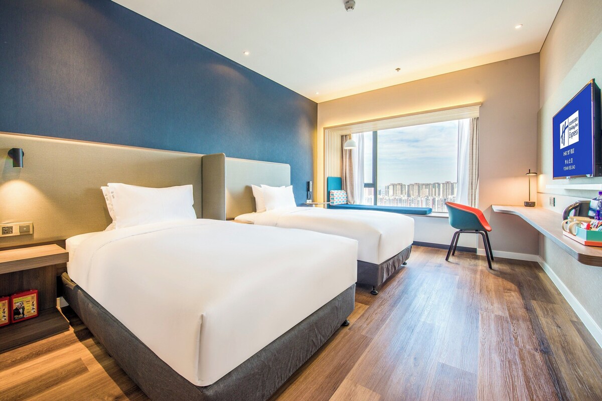 A hotel room with two beds, a desk, a chair, and a large window with a city view.