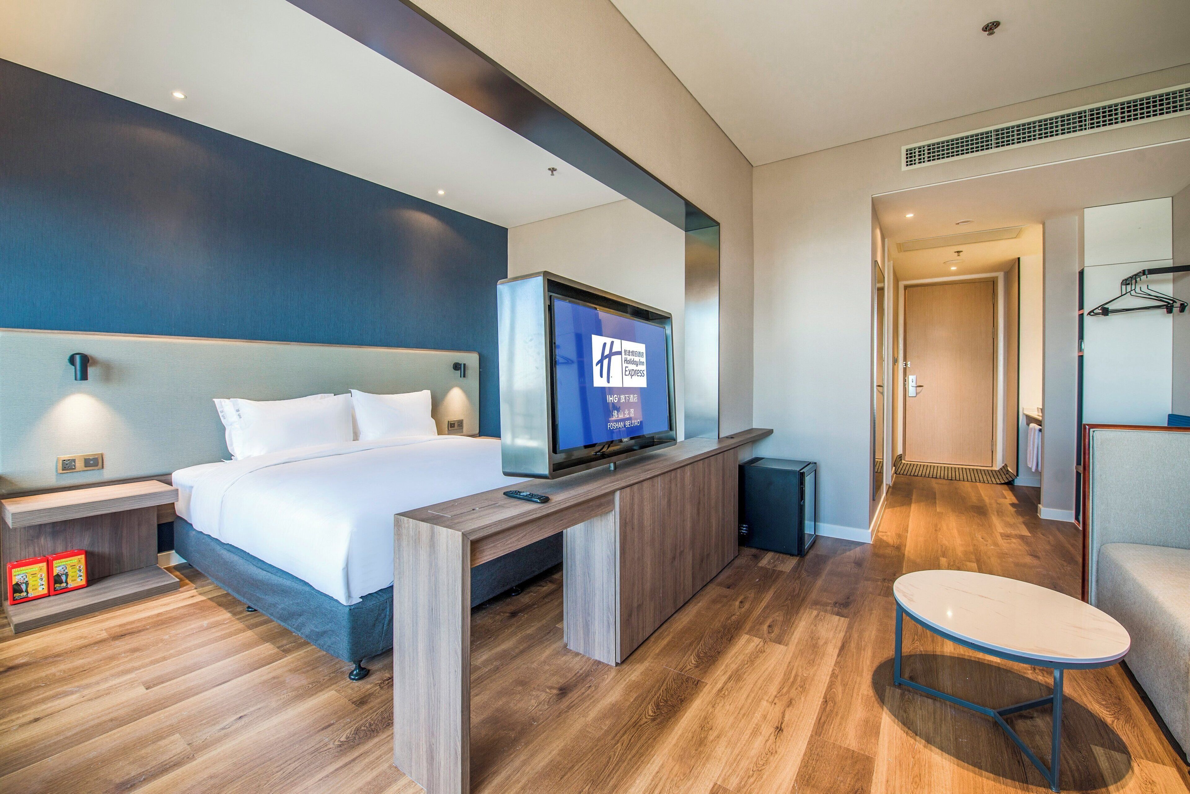A hotel room with a bed, a TV mounted on a wooden console, a small table, and a chair.