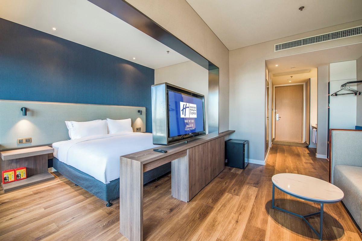 A hotel room with a bed, a TV mounted on a wooden console, a small table, and a chair.