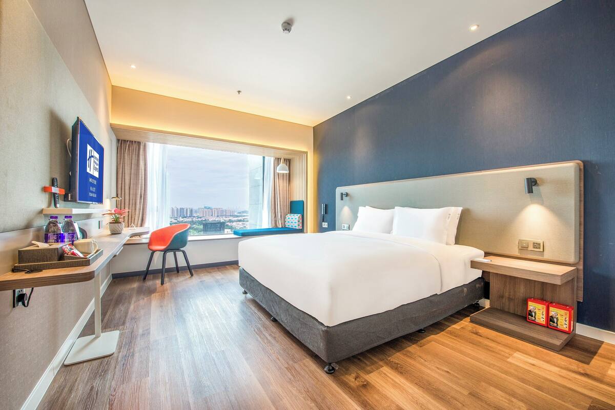 A hotel room with a large bed, a desk with a chair, a TV, and a window with a city view.