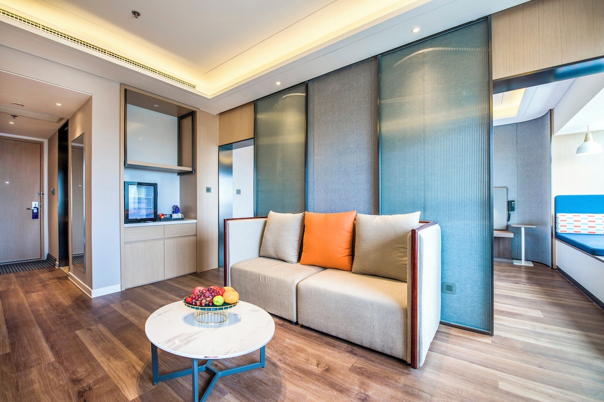A modern hotel room with a sofa, a round table with fruit, a kitchenette, and a large window.
