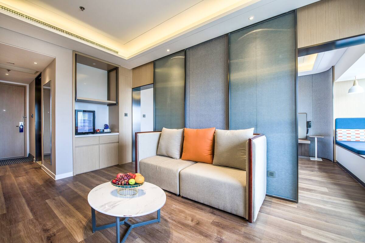 A modern hotel room with a sofa, a round table with fruit, and a view of the city.