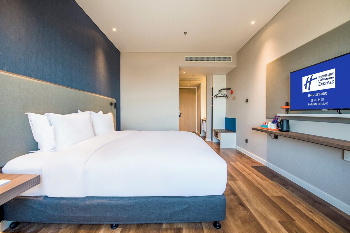 A modern hotel room with a large bed, a flat-screen TV mounted on the wall, and a wooden floor.