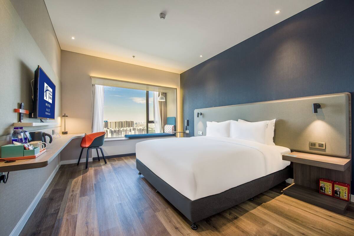 A modern hotel room with a large bed, a desk with a chair, a TV, and a window with a city view.