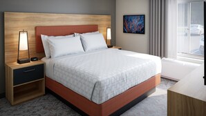 A hotel room with a large bed, two bedside tables with lamps, a small shelf, a painting on the wall, and a view of parked cars through the window. - Candlewood Suites Midland South I 20 by IHG (Midland)