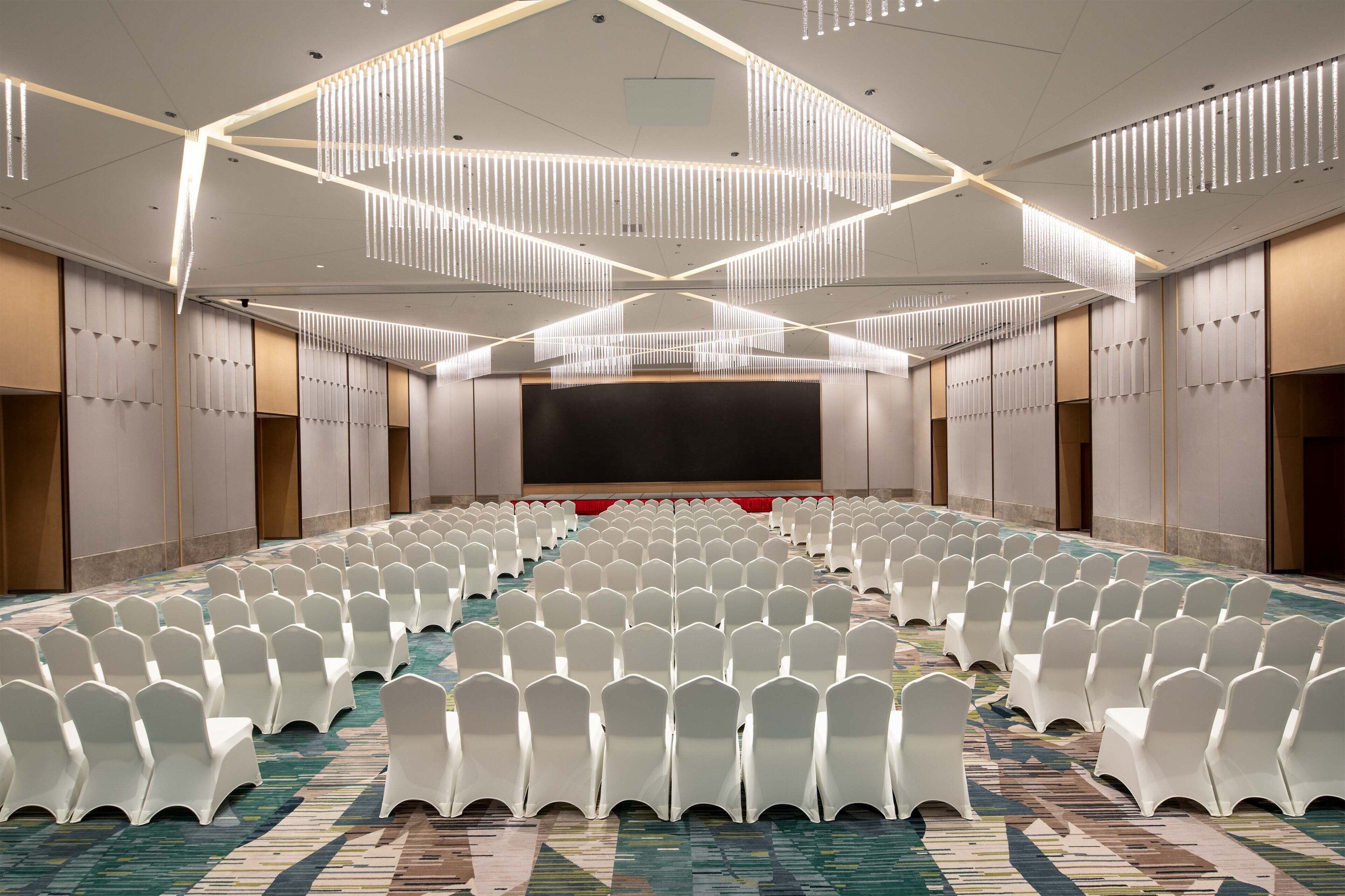 A conference room with rows of white chairs, a large screen, and modern lighting fixtures.