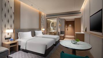 Una habitación de hotel moderna con dos camas, una zona de estar con una mesa redonda, un televisor y un baño con cabina de ducha de vidrio.