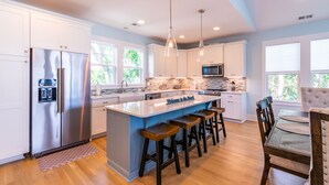 A modern kitchen with white cabinets, stainless steel appliances, a central island with bar stools, pendant lighting, and large windows. - Brand NEW! | Luxury Townhome | 2 Master Suites | Elevator | Pool and ocean view (Kure Beach)