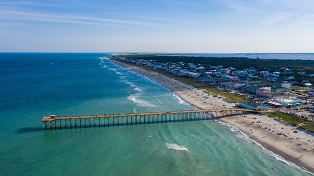 An aerial view of a coastal area with a pier extending into the ocean, a beach with people, and a row of buildings along the shore. - Brand NEW! | Luxury Townhome | 2 Master Suites | Elevator | Pool and ocean view (Kure Beach)