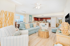 A spacious living room with a sofa, armchairs, and a coffee table. A kitchen is visible in the background with wooden cabinets and a microwave.