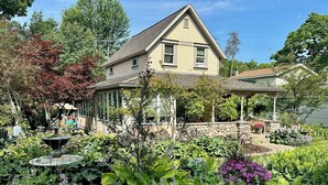 A house with a covered porch, surrounded by a garden with various plants and a fountain. - Historical home, the Burnett Nestt in the heart of Lake Ann  (Lake Ann)
