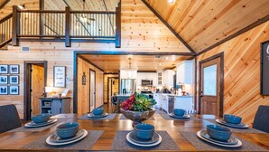 Interior de madera con comedor listo para seis personas, cocina al fondo y una escalera que lleva a un nivel superior.
