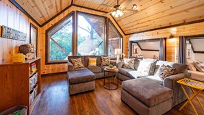 Un salón acogedor, con muebles de madera, ventanas amplias, un ventilador de techo y una pequeña mesa con un tablero de ajedrez.