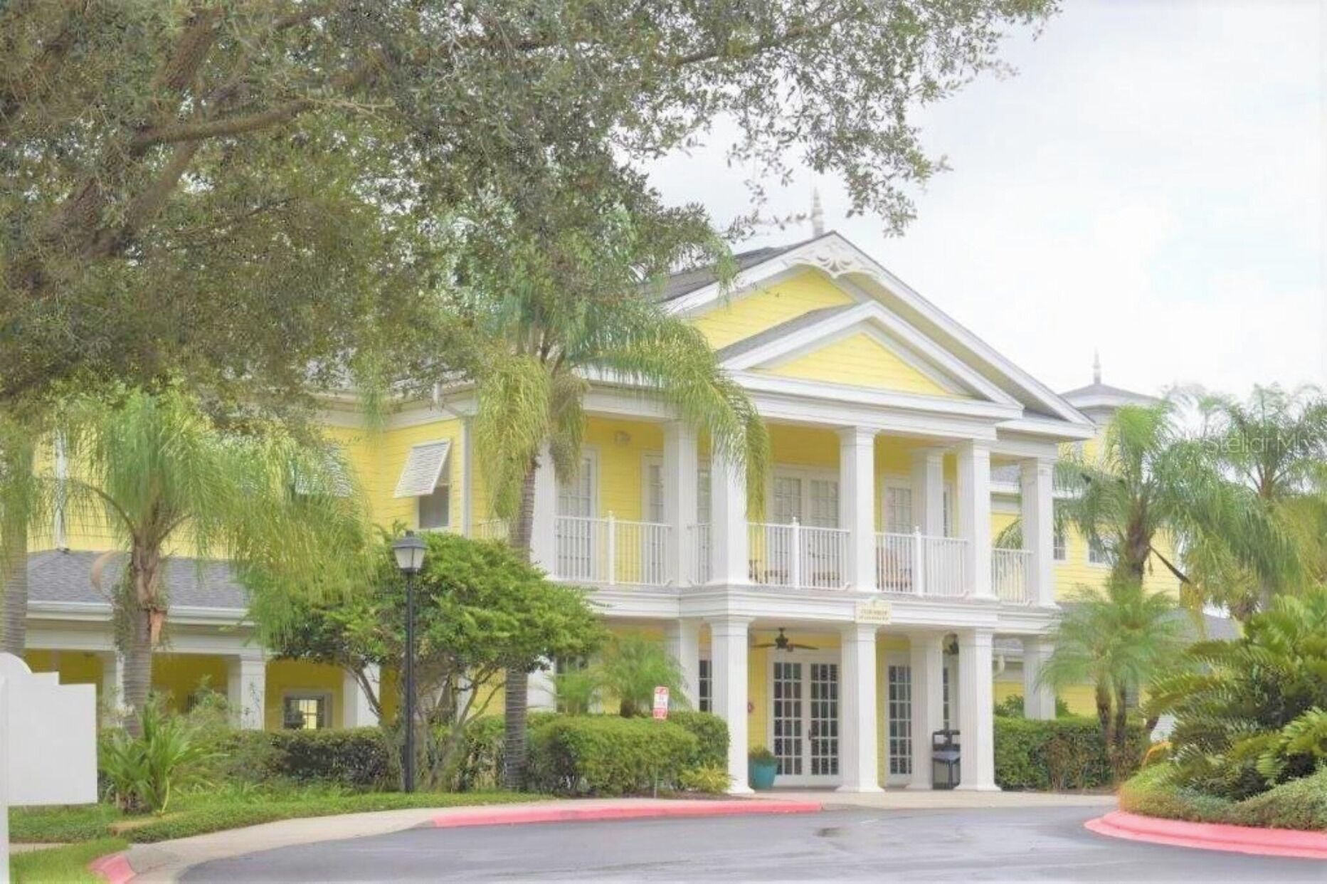 A yellow building with a balcony and white columns, surrounded by greenery.