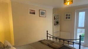 A bedroom with a bed, a bedside lamp, a window, and a shelf with books and a plant. - Cute Convenient and close to the beech. (Brighton)