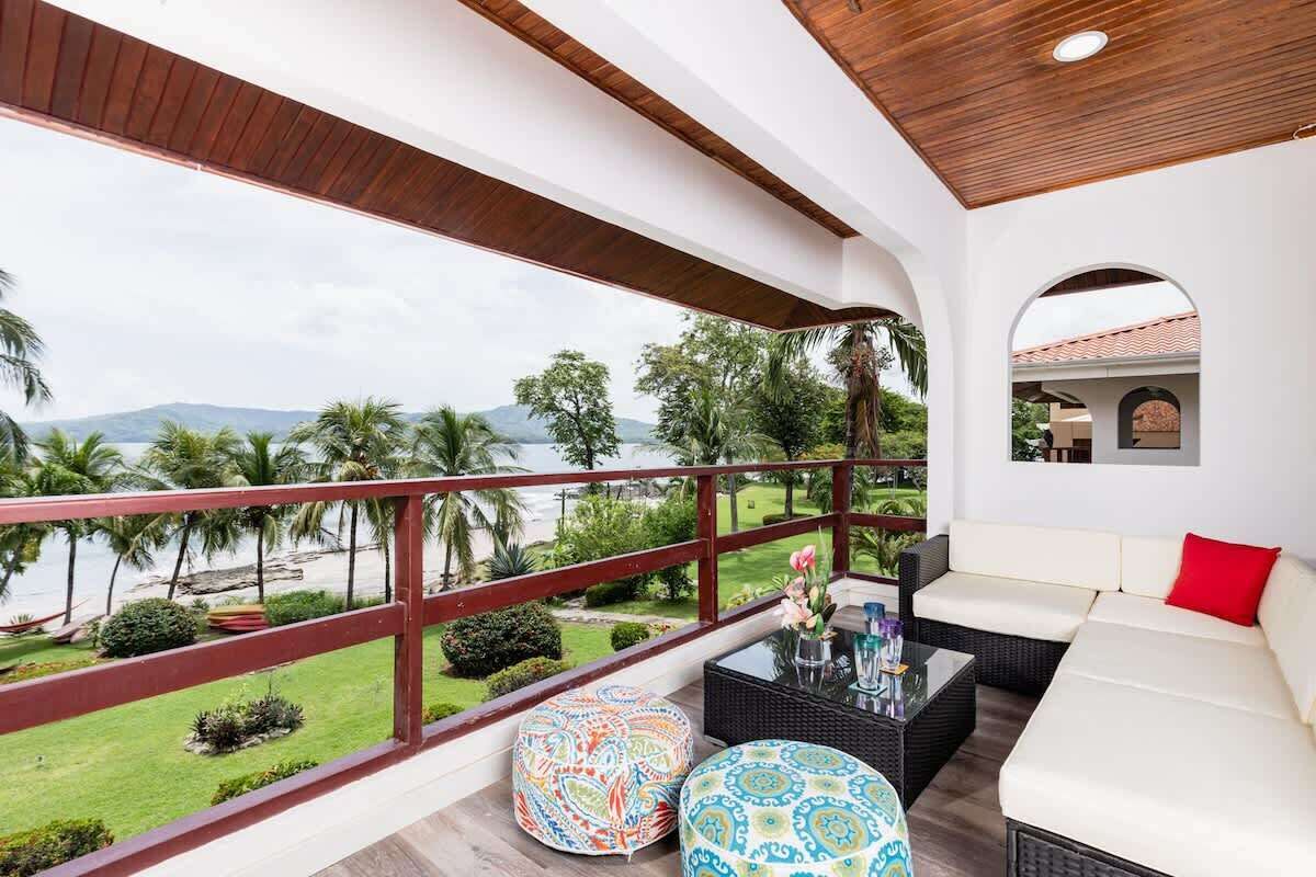 Uma varanda com vista para a praia e palmeiras, um sofá branco e pufes estampados.