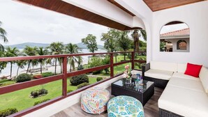 A balcony with a view of the beach and palm trees, a white sofa, and patterned ottomans.