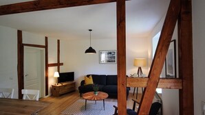 Una sala de estar con piso de madera, un sofá negro, una mesa redonda para café, un televisor sobre un mueble de madera y una lámpara en una mesita auxiliar.