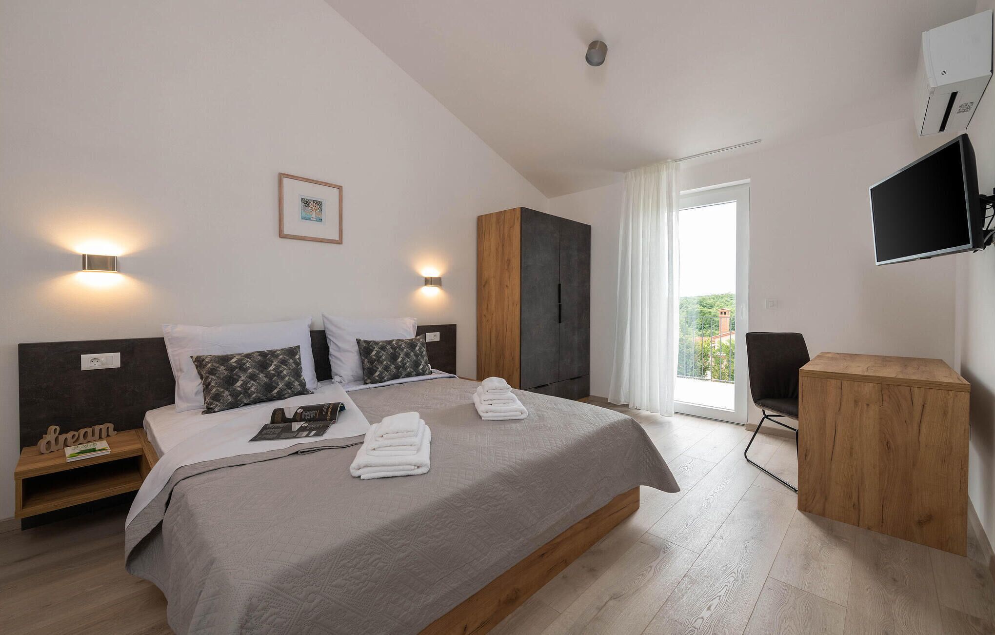 A modern hotel room with a large bed, two bedside tables, a wardrobe, a TV, and a view of the outdoors.