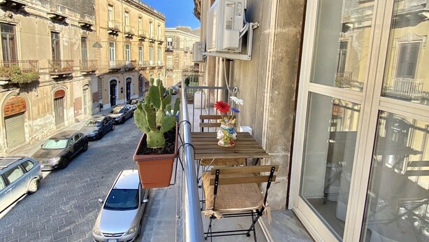 A narrow street with parked cars, a cactus planter, and a small table with flowers on a balcony.