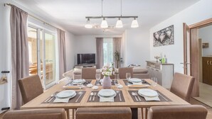 A dining area with a wooden table set for four, surrounded by chairs. A television is mounted on the wall, and there is a plant on the windowsill. - Beautiful home in Orihi (Orihi)