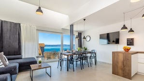 A modern living room with a sofa, dining table, and chairs, a kitchen area with a wooden counter, and a large window offering a view of the sea. - Luxurious home in Njivice (Njivice)