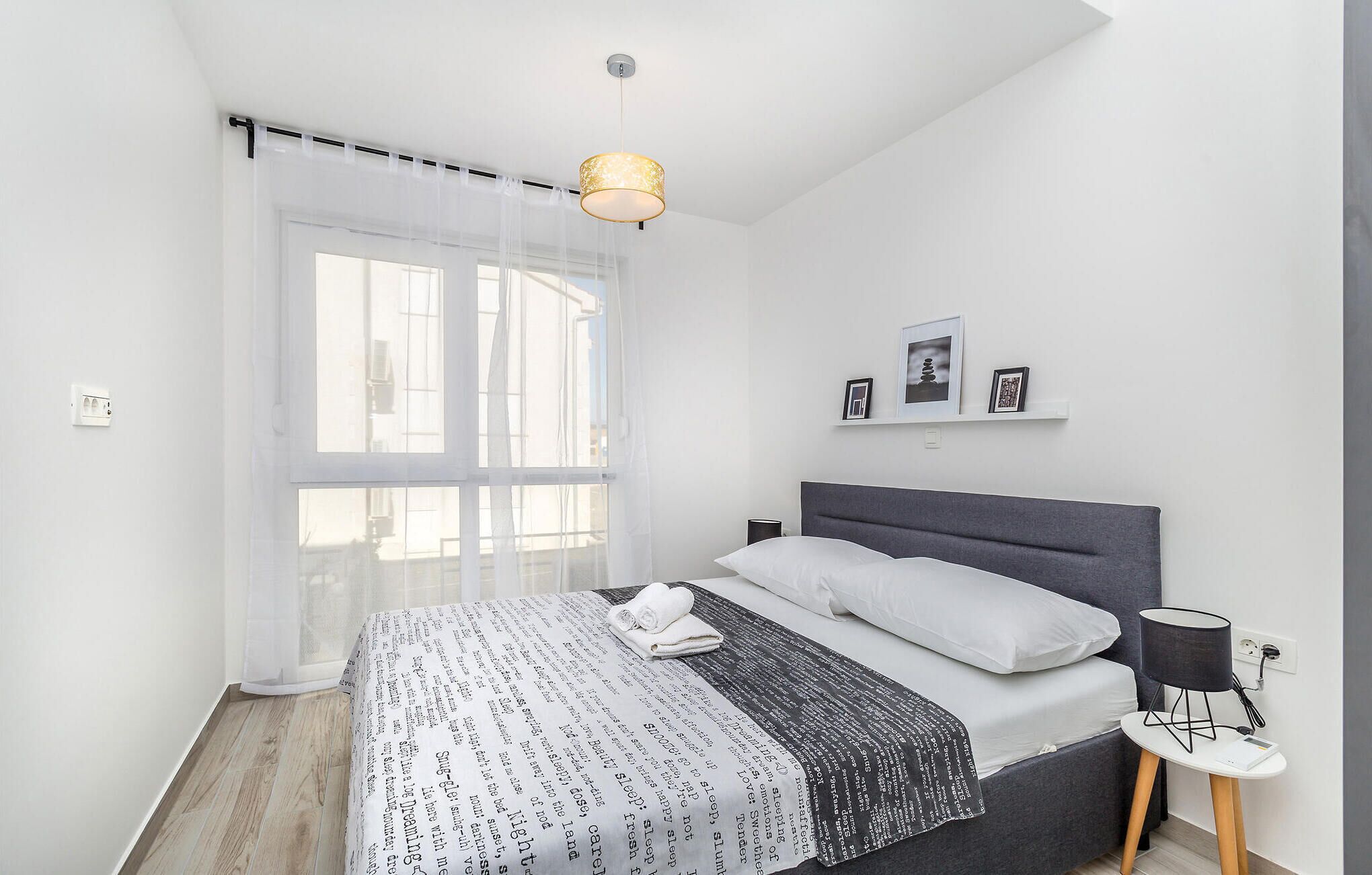 Ein modernes Schlafzimmer mit großem Fenster, einem Bett mit grauem Kopfteil, einem Nachttisch mit Lampe und einem Wandregal mit Bildern.