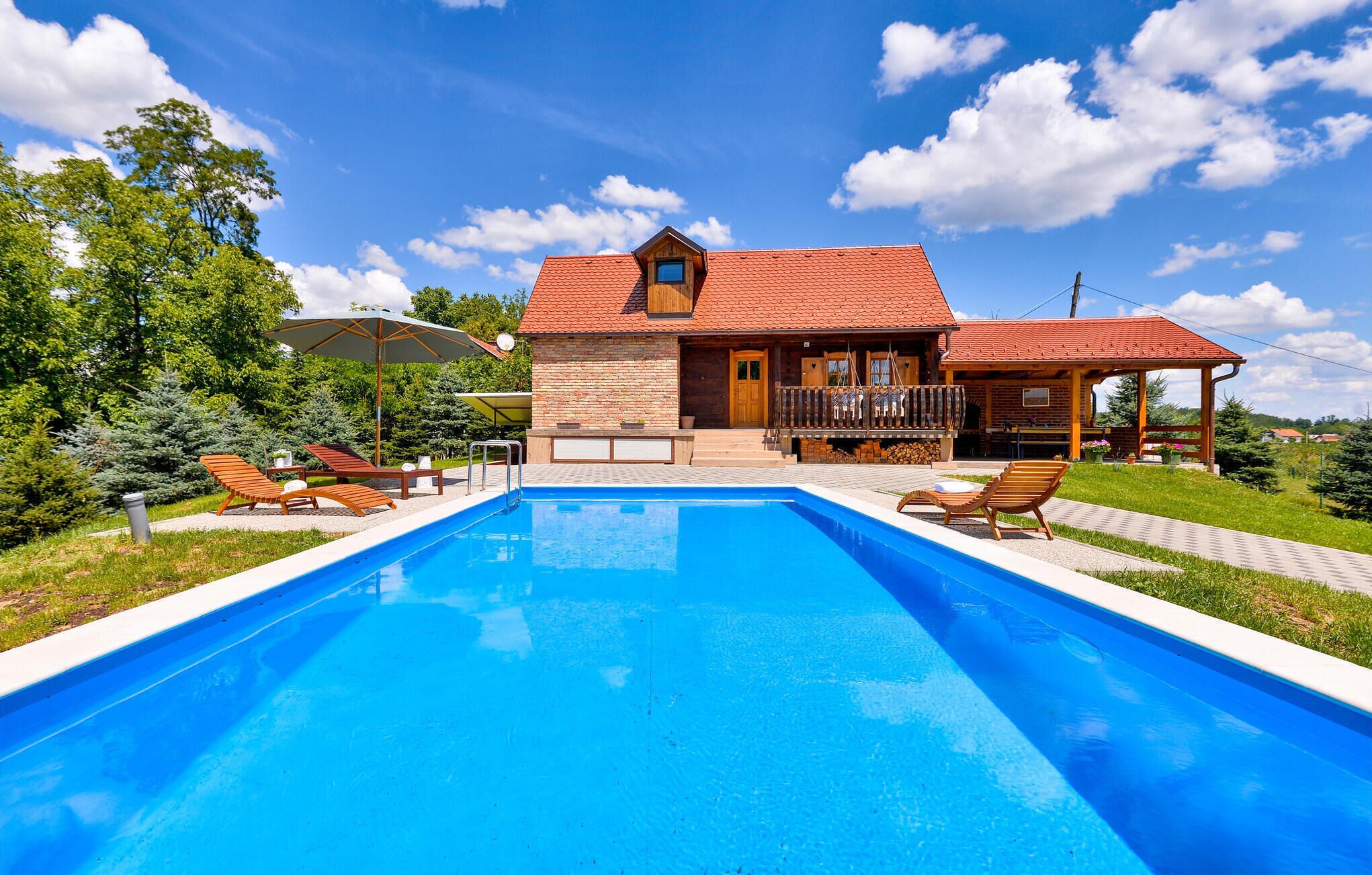 A swimming pool with lounge chairs, a wooden house with a red roof, and a covered patio area.