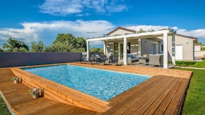 A modern outdoor pool area with a wooden deck, lounge chairs, and a covered seating area.