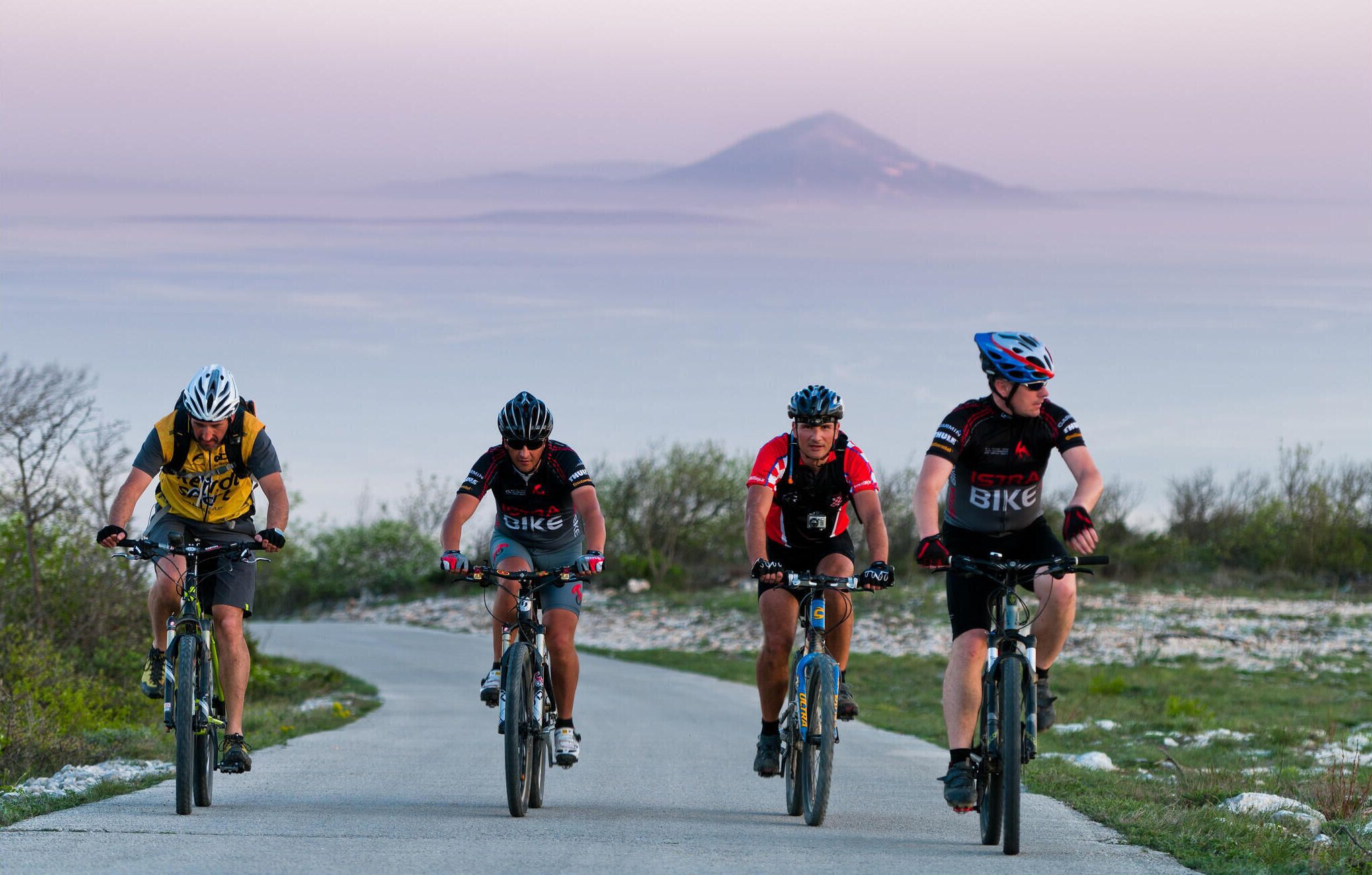 Quattro ciclisti su una strada asfaltata con montagne sullo sfondo.