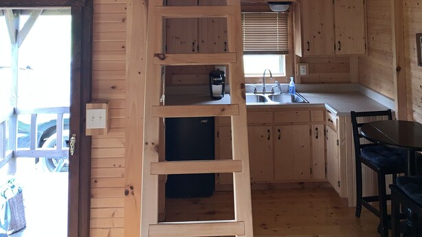 A wooden cabin with a loft bedroom, a kitchen with wooden cabinets, a ladder leading to the loft, and a ceiling fan. - Cozy Cabin with private back yard, located 5 min away from Dale Hollow Lake (Albany)