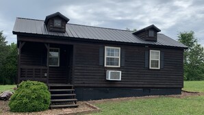 A small, dark wooden cabin with a metal roof, a front porch, and a small garden area. - Cozy Cabin with private back yard, located 5 min away from Dale Hollow Lake (Albany)