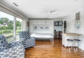 A bedroom with a bed, a desk with chairs, a ceiling fan, and a large window.