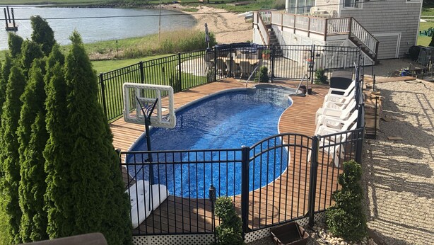 Une piscine avec un panier de basketball, entourée d’une terrasse en bois et d’une clôture en métal.