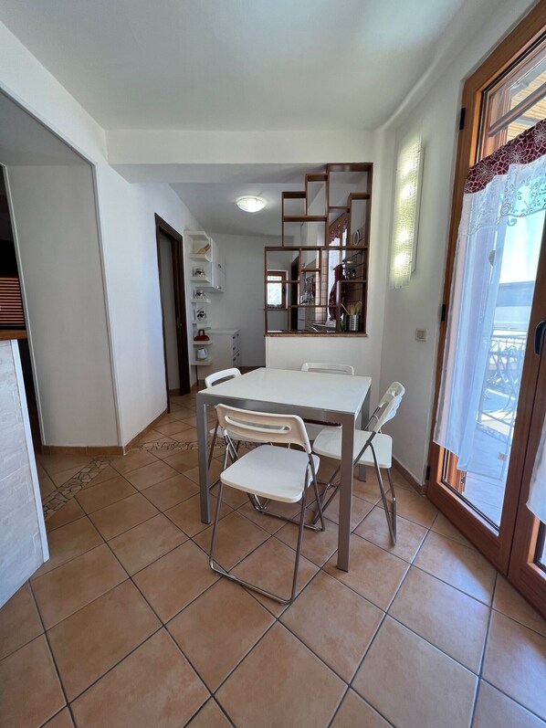 A dining area with a white table and chairs, a kitchen area with shelves and cabinets, and a sliding glass door leading to an outdoor balcony.