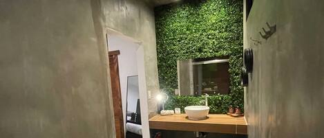 Un baño compacto con diseño moderno, que incluye un inodoro blanco, un tocador de madera con espejo y un detalle de pared verde.