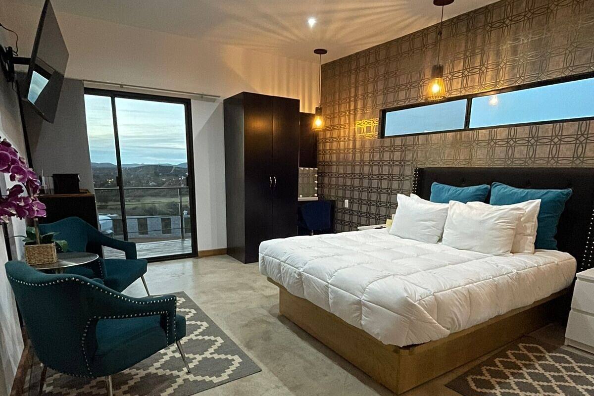 A modern bedroom with a large bed, a desk, and a chair. There is a view of the city through the window.