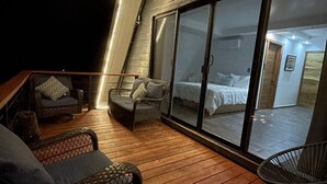 Un balcón acogedor con mobiliario de mimbre, piso de madera y una puerta corrediza de vidrio que da a un dormitorio con cama y mesita de noche.