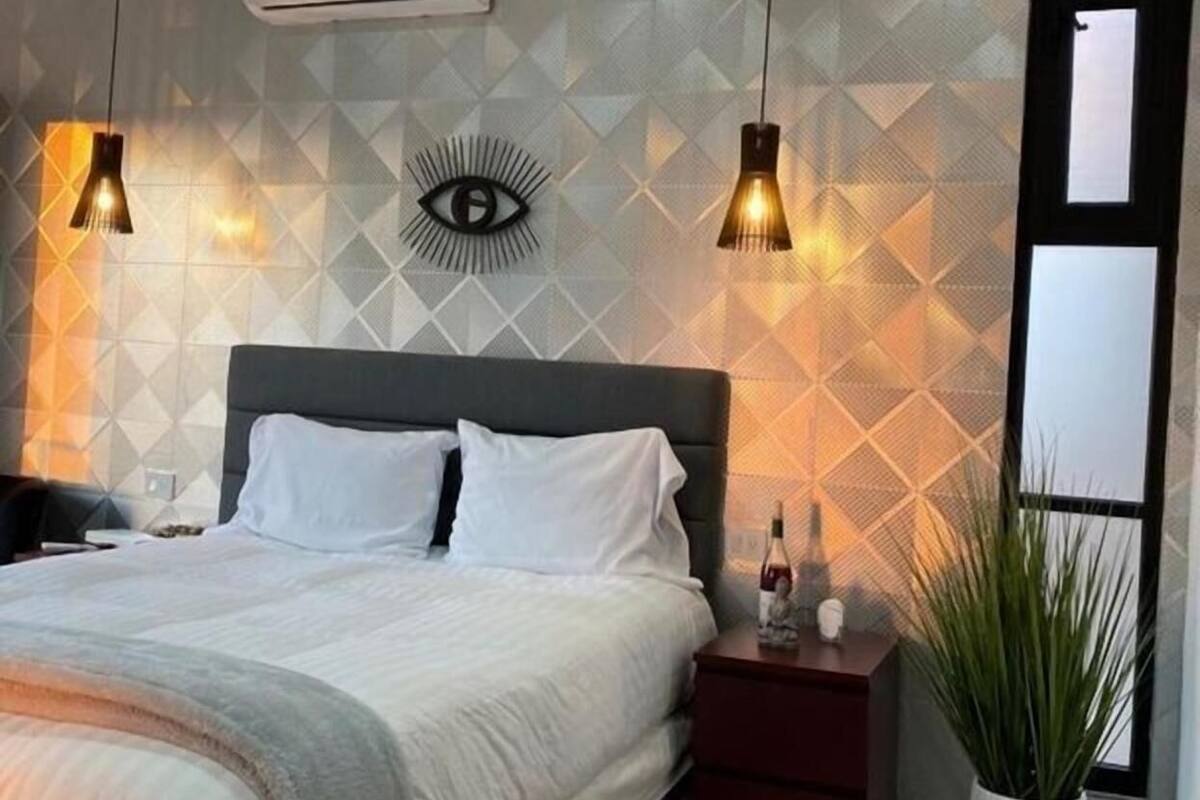 A modern bedroom with a bed, two pendant lights, a wall-mounted air conditioner, a geometric-patterned wall, and a potted plant.