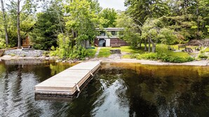 A lakeside cabin with a dock extending into the water, surrounded by dense trees and a well-maintained lawn. - Lakefront Muskoka Retreat 4 Bedroom Cottage  (MacTier)