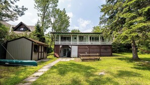 A two-story house with a balcony, a covered porch, and a wooden bench in the yard. - Lakefront Muskoka Retreat 4 Bedroom Cottage  (MacTier)