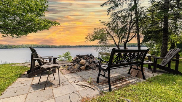 A lakeside seating area with a bench and chairs, a fire pit, and a view of the lake and sunset. - Lakefront Muskoka Retreat 4 Bedroom Cottage  (MacTier)