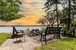 A lakeside seating area with a bench and chairs, a fire pit, and a view of the lake and sunset. - Lakefront Muskoka Retreat 4 Bedroom Cottage  (MacTier)