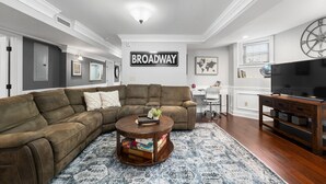 Un salon avec un canapé d’angle, une table basse avec des livres, un téléviseur et un mur orné d’un panneau indiquant « BROADWAY ».
