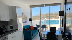 Eine moderne Küche mit weißen Schränken, einer Mikrowelle und einem Herd. Ein blaues Sofa mit orangefarbenen Kissen, ein Esstisch mit Stühlen und Blick auf das Meer durch große Fenster.