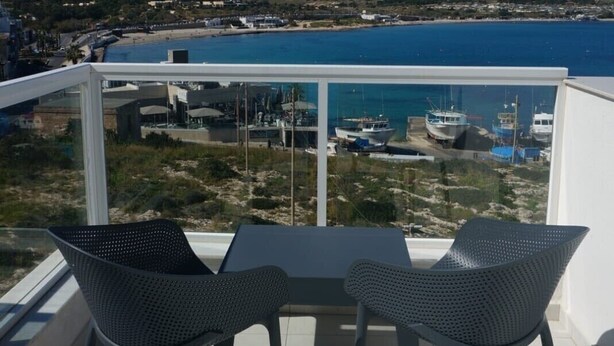 Ein Balkon mit zwei Stühlen und einem Tisch, der auf eine Küstenlandschaft mit Booten und einem Strand blickt.