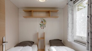 A small, clean room with two single beds, a wooden shelf above the beds, and a window with curtains.